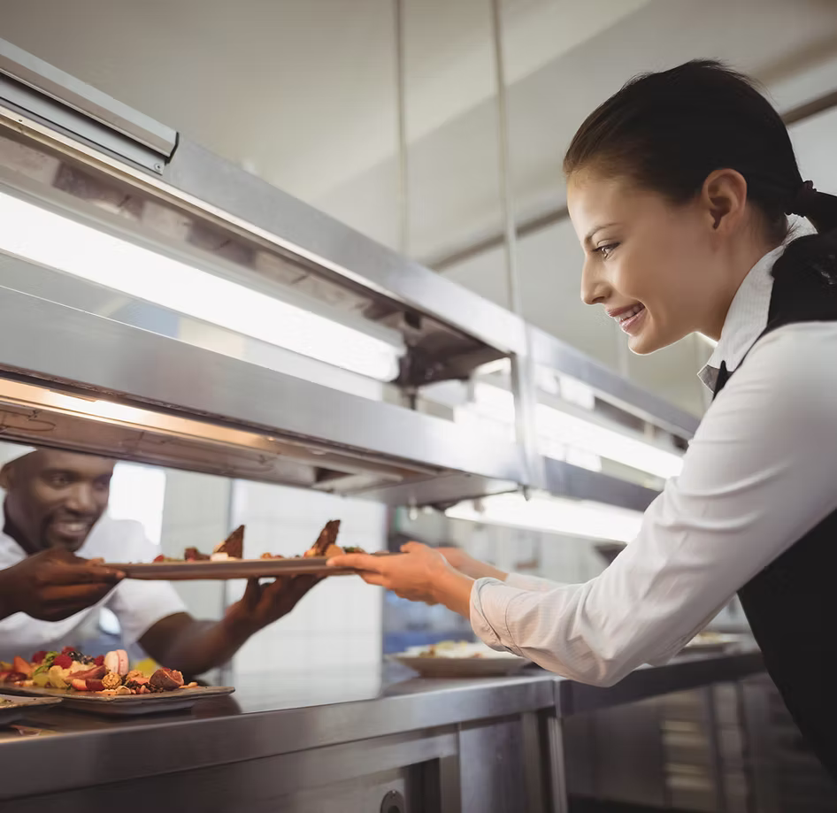 Two chefs in a kitchen, one serving food from an oven, the other holding a platter.