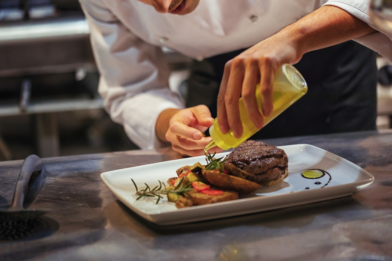 Chef garnishing a dish with a bottle of oil on a professional kitchen counter.
